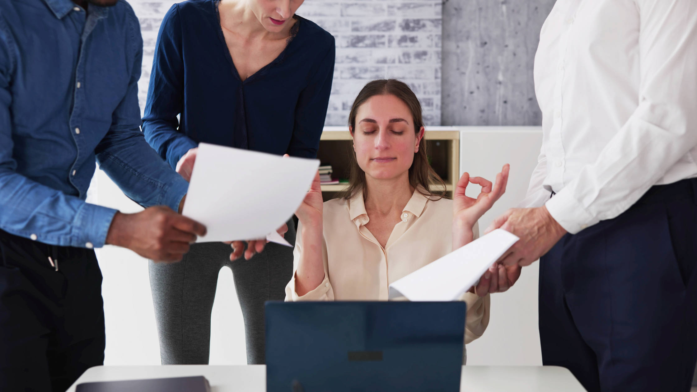 Female executive sitting at her desk making meditation hand gestures while surrounded by colleagues asking for help, symbolizing stress management in leadership.